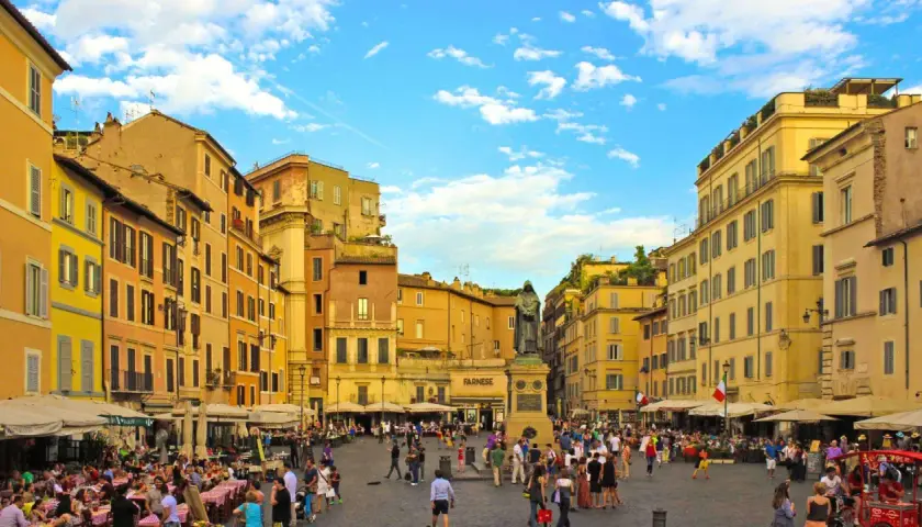 Place animée de Campo de' Fiori à Rome, avec des terrasses de café, des bâtiments colorés et une statue au centre.