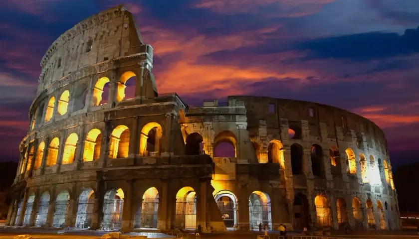 Le Colisée de nuit, illuminé par des lumières chaudes, se dresse sous un ciel aux couleurs flamboyantes.