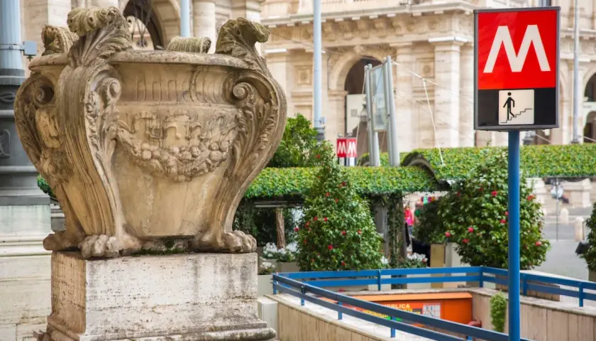 Un signe du **metro vatican** indique l'entrée d'une station, à côté d'une urne antique et de verdure luxuriante.