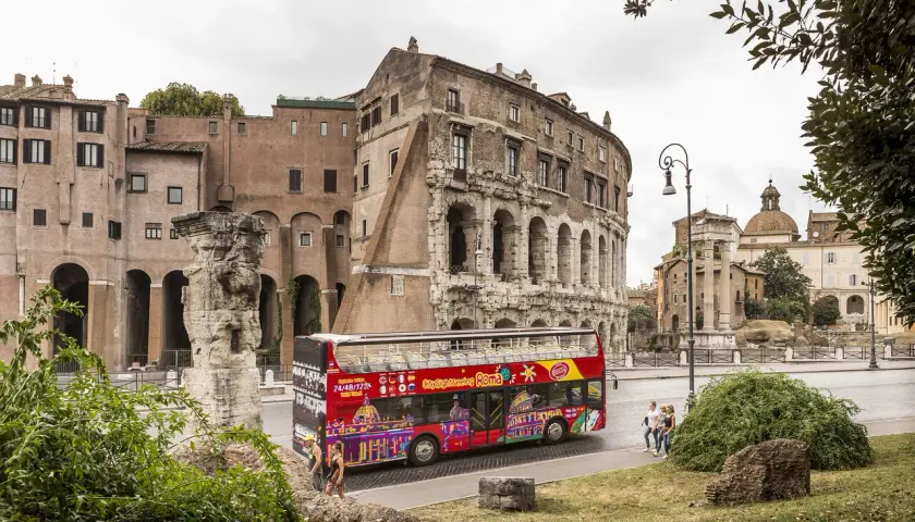 Un bus touristique rouge vif, "Citysightseeing Roma", passe devant des ruines antiques et des bâtiments romains.
