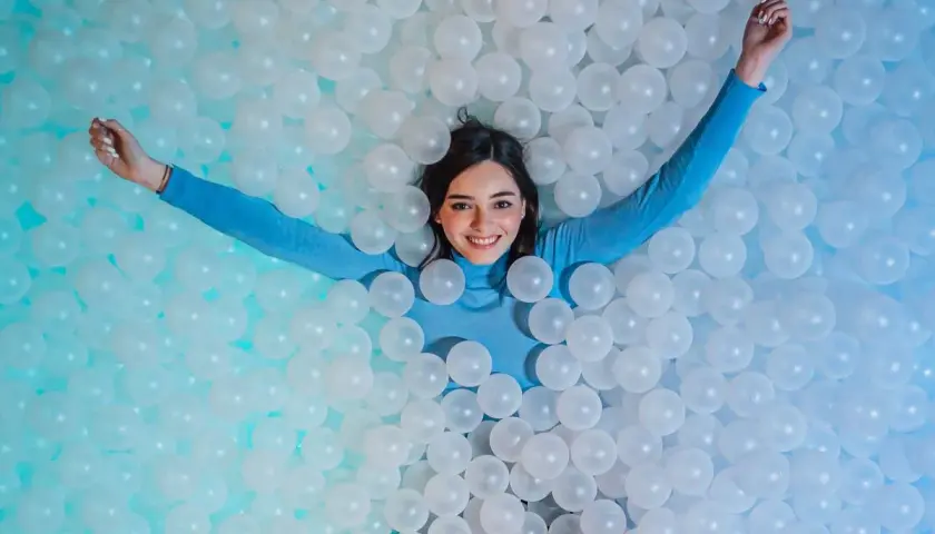 Une jeune femme souriante se détend dans une piscine de balles blanches, un clin d'œil ludique à l'ikono Rome.