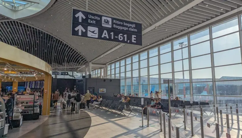 Intérieur lumineux d'un aéroport, avec des panneaux indiquant "Uscita" et "Ritiro bagagli". Des voyageurs attendent près des grandes baies vitrées, prêts pour leur vol au départ de l'aéroport Rome centre ville.