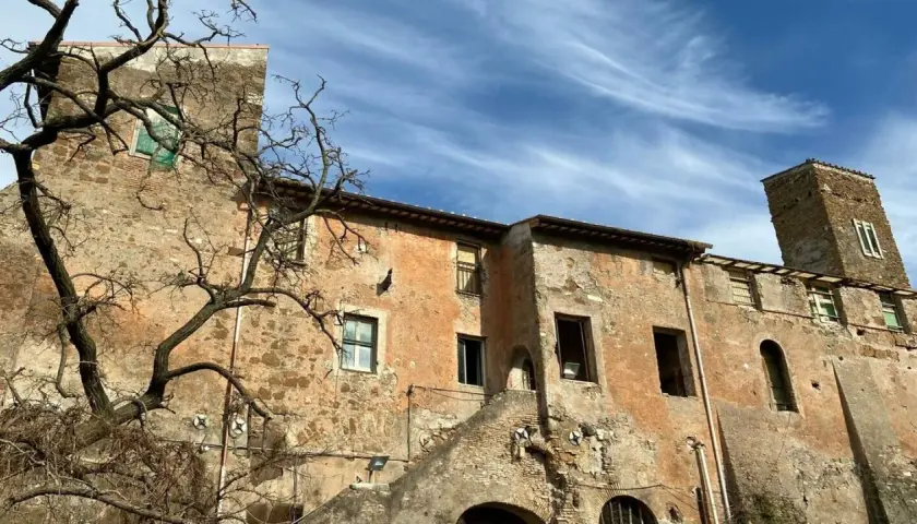 Bâtiments anciens et décrépits, avec une tour de briques penchée, sous un ciel bleu et nuageux. Une promenade sur la Via Appia à pied révèle ces ruines.