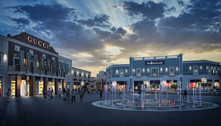 Un soir à l'outlet Sicile, les fontaines illuminées dansent sous un ciel dramatique, devant les boutiques Gucci et Polo Ralph Lauren.