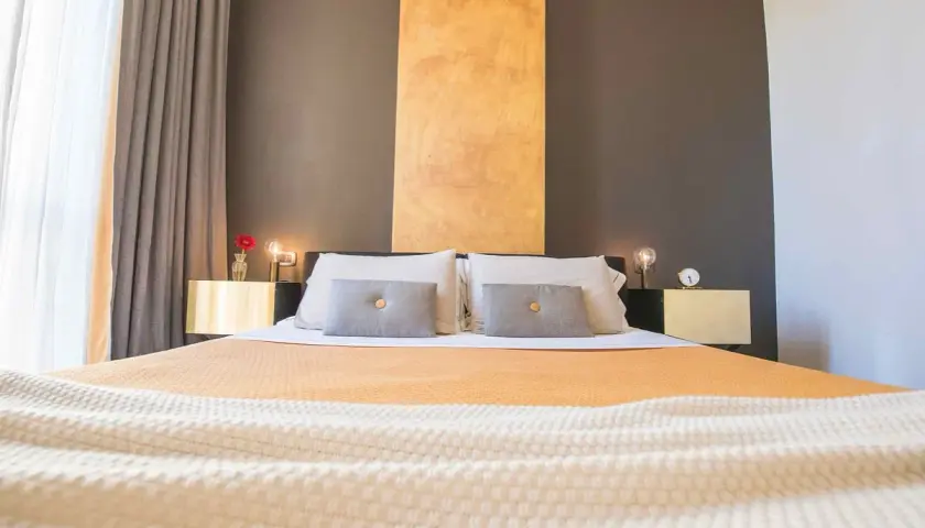 Lit et tables de chevet dorés dans une chambre d'hôte Italie du Nord. Rideaux gris et blancs, mur anthracite et ocre.