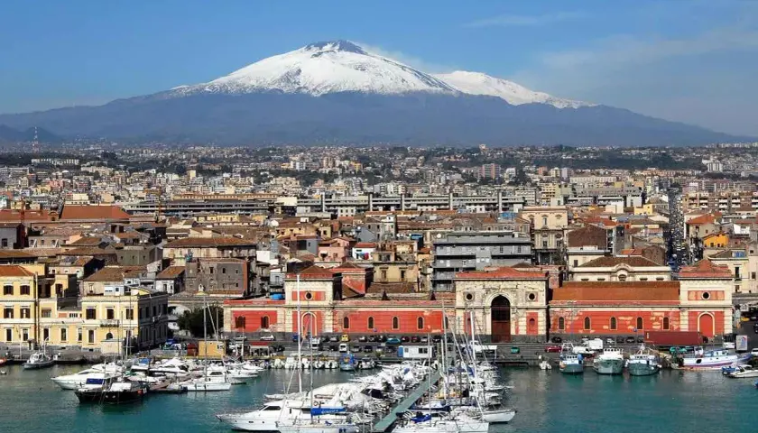 Vue panoramique de la ville de Catania, Sicile, avec le mont Etna enneigé en arrière-plan et un port rempli de voiliers.