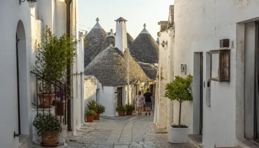 Ruelle pavée bordée de maisons blanches aux toits coniques typiques des Pouilles à éviter, avec un couple se promenant.