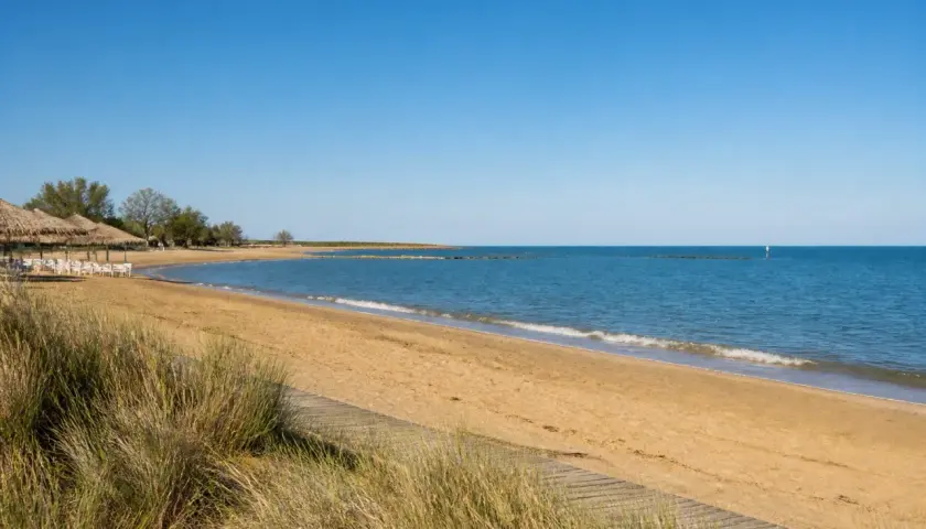Plage de sable doré à Caorle, avec des parasols de chaume, des chaises blanches et la mer bleue sous un ciel clair.