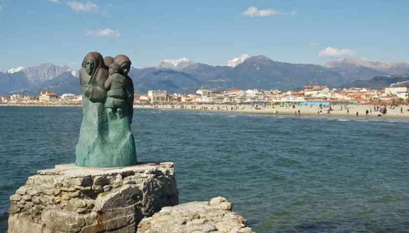 Sculpture de famille sur un rocher, face à la mer et à la ville de via Reggio, avec des montagnes enneigées en arrière-plan.