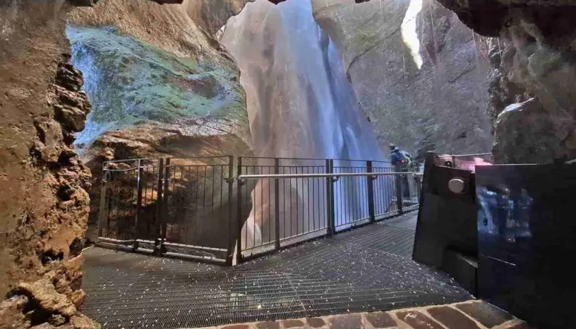Vue depuis une grotte sur la cascade du Varone, avec une passerelle et un visiteur admirant la chute d'eau.