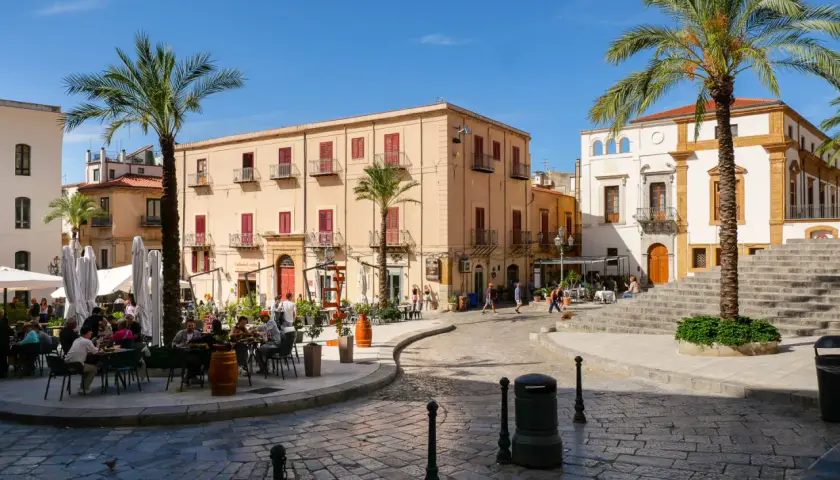 Place animée de Cefalù avec des terrasses de café, des palmiers et des bâtiments historiques sous un ciel bleu.
