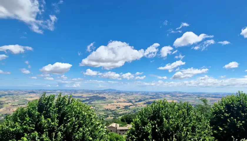Vue panoramique depuis Cingoli, avec des champs ondulants et un ciel bleu parsemé de nuages blancs.