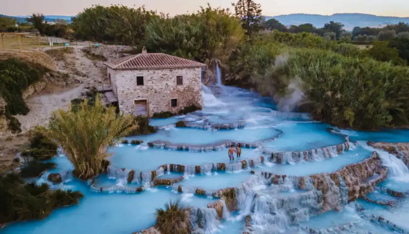 Casciana Terme : un couple se promène dans les cascades d'eau thermale bleue, près d'une vieille bâtisse en pierre.