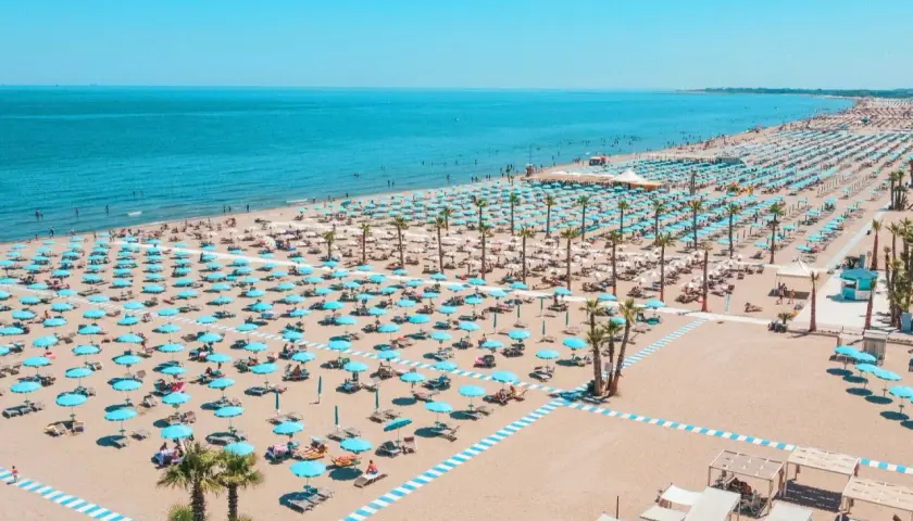 Plage de Chioggia, Italie, avec des rangées infinies de parasols bleus et blancs, des chaises longues et des palmiers sous un ciel bleu éclatant.
