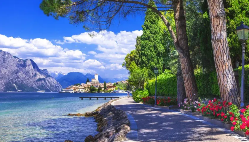 Promenade ensoleillée au bord du lac de Garde, avec des montagnes majestueuses en arrière-plan et un village pittoresque.