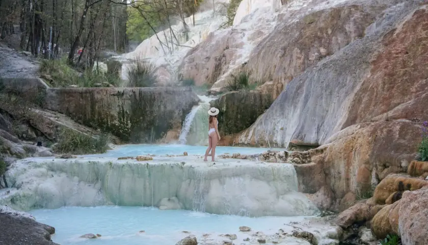 Une femme en maillot de bain blanc et chapeau de paille admire les cascades et les bassins turquoise des Bagni San Filippo, un paysage naturel spectaculaire.