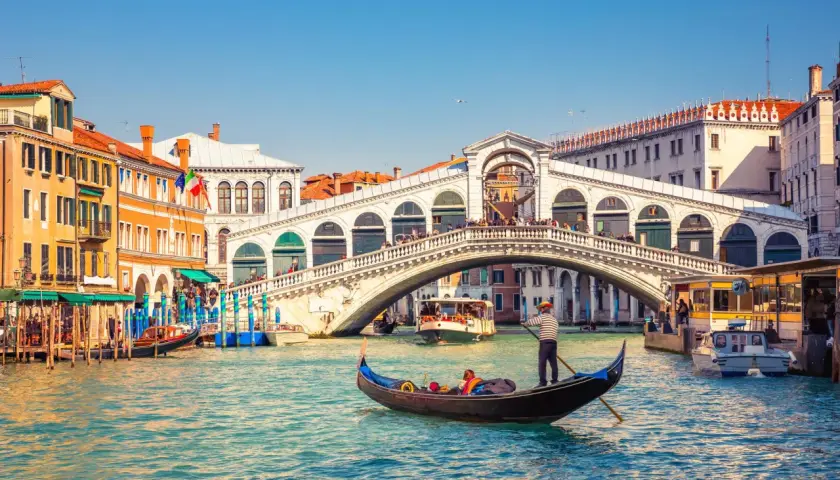 Gondole naviguant sur le Grand Canal, sous le célèbre pont du Rialto à Venise. L'eau turquoise reflète les façades colorées des bâtiments.