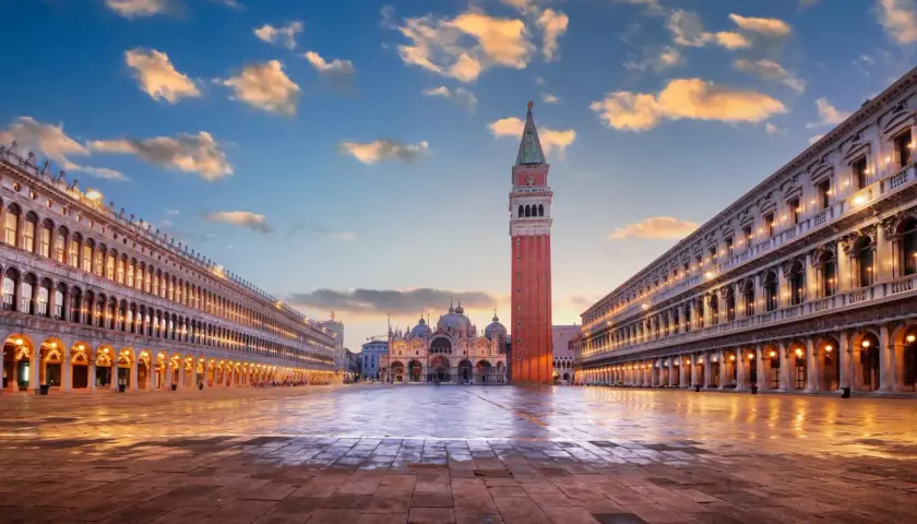 Le **place Saint-Marc à Venise** s'illumine au crépuscule. La basilique et le campanile se dressent majestueusement sous un ciel nuageux.