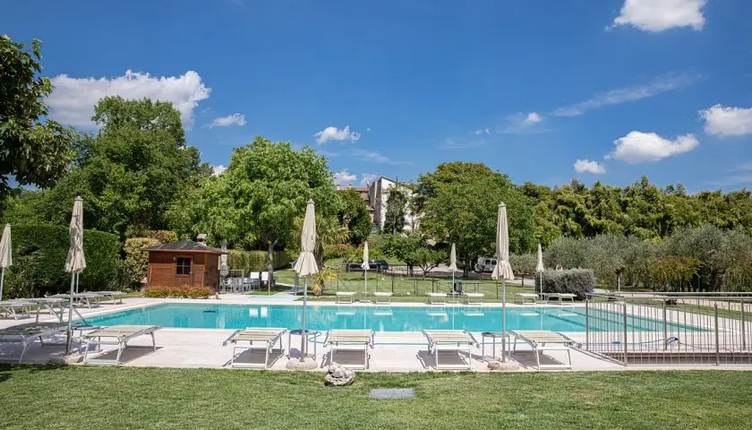 Piscine d'agritourisme au Lac de Garde, entourée de verdure, avec chaises longues et parasols sous un ciel bleu.
