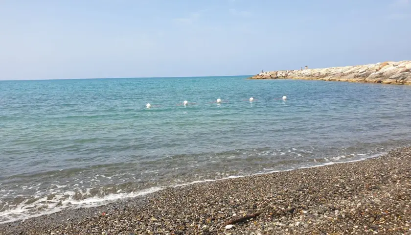 Plage de galets à Cecina Mare, avec des bouées blanches marquant la zone de baignade et une jetée rocheuse au loin.