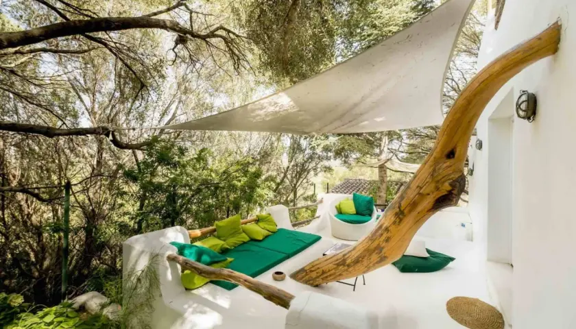Terrasse du Hotel Su Gologone avec mobilier blanc et coussins verts, sous une voile d'ombrage et entourée de verdure.