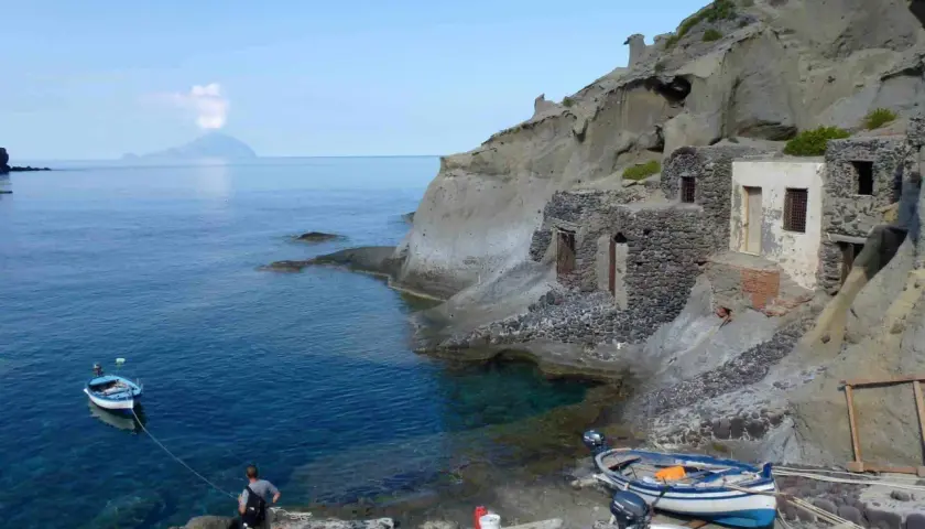 Sur l'île Salina, un homme amarre un bateau près de vieilles maisons de pêcheurs en pierre, avec une île volcanique au loin.
