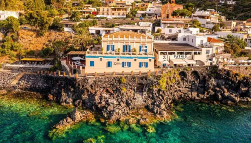 Vue d'un gîte en Sicile, perché sur une falaise rocheuse surplombant une mer turquoise. Bâtiments blancs et ocres, végétation luxuriante.