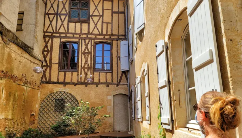 Cour intérieure avec colombages et volets blancs. Une femme regarde la scène, comme si elle découvrait un coin charmant de Fleurance, Italie.