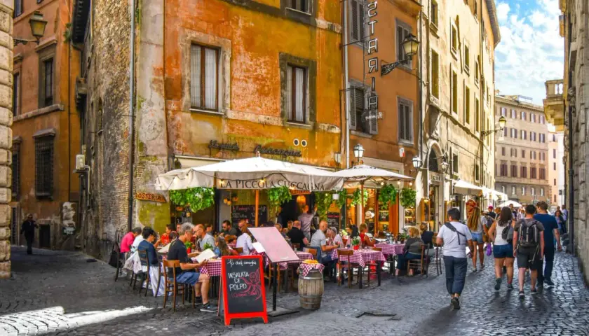 Une trattoria animée à Rome, avec des tables dressées sur la rue pavée. Des gens savourent un repas sous les parasols, profitant de l'ambiance romaine.