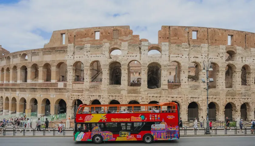 Un bus touristique rouge vif circule devant le Colisée à Rome, transportant des visiteurs pour une visite de la ville.