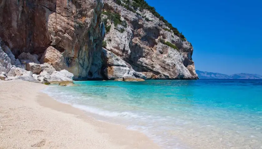 Plage de sable fin bordée par une eau turquoise cristalline, avec une falaise imposante en arrière-plan. Un coin de paradis sur la côte smeralda sardaigne.