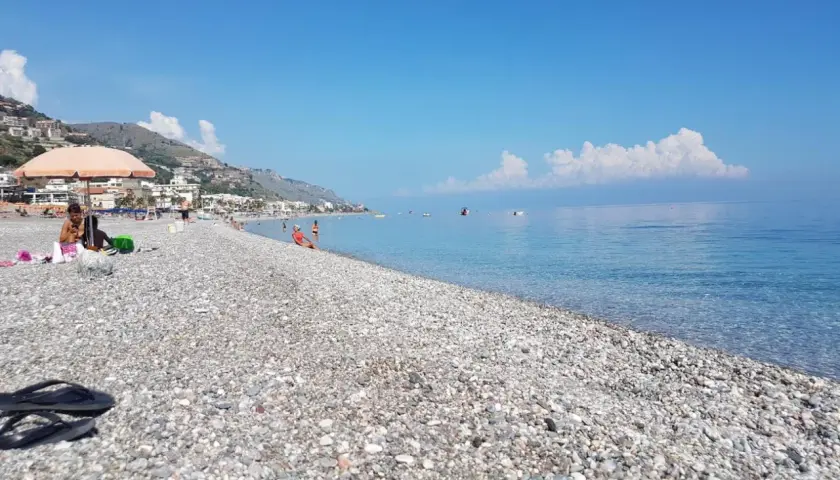 Plage de galets à Letojanni, Sicile. Des baigneurs profitent du soleil sous un parasol, tandis que la mer d'un bleu profond s'étend à l'horizon.