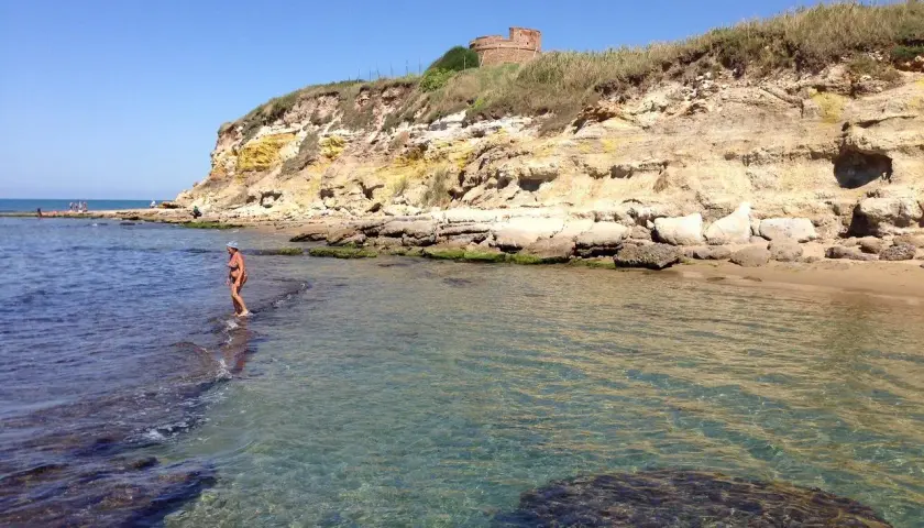 Une femme en bikini profite de la mer limpide près d'une falaise rocheuse à Anzio, avec une tour ancienne en arrière-plan.