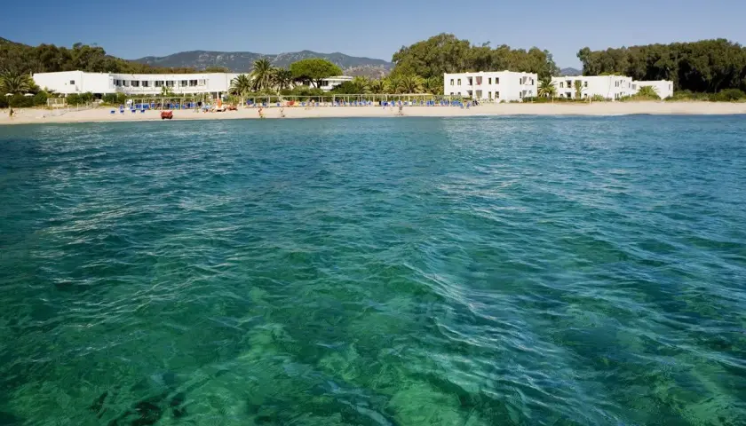 Eaux turquoise cristallines devant une plage de sable fin à Pula, Sardaigne, avec des hôtels blancs et des palmiers.