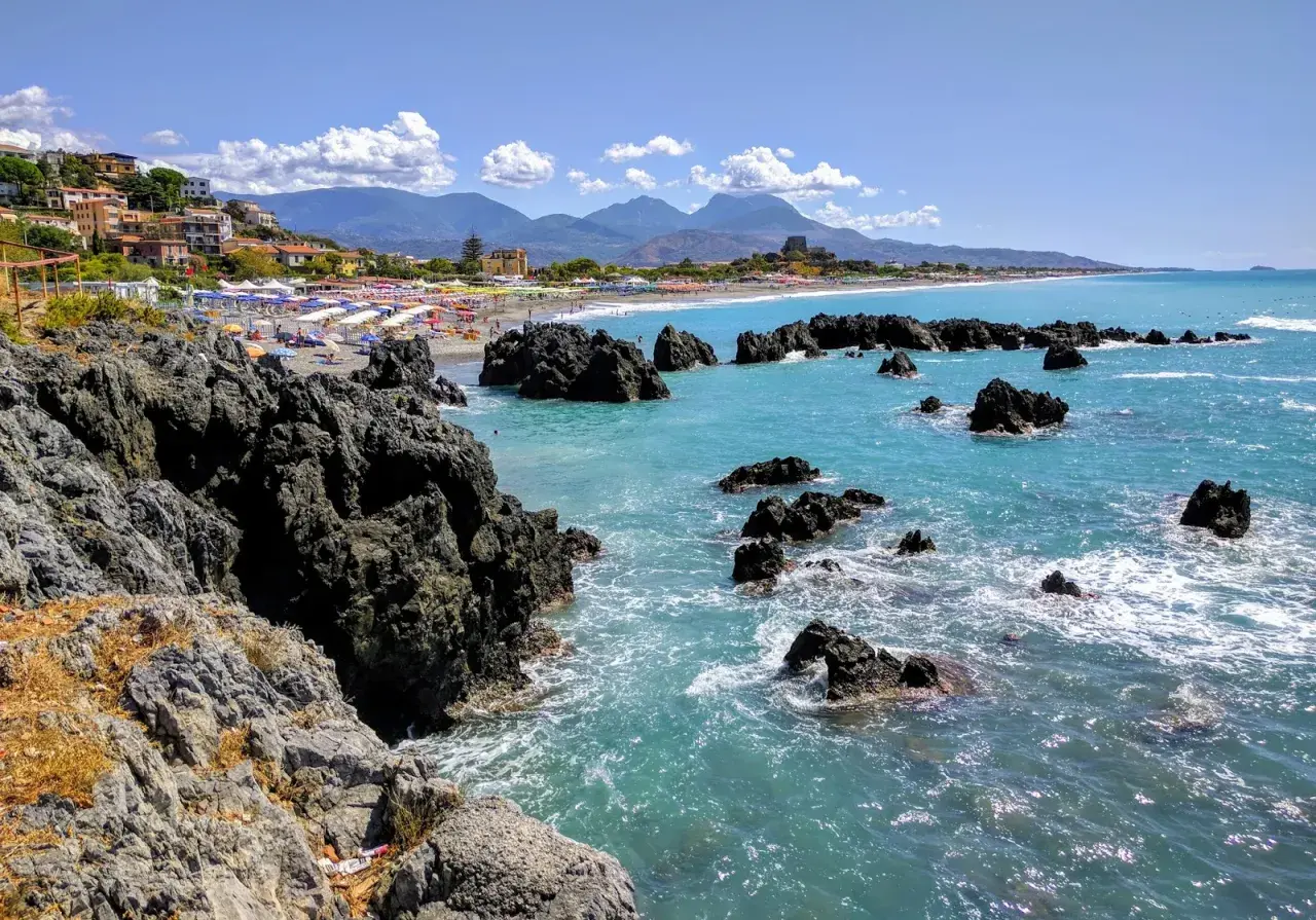 Vue d'une plage animée avec des parasols colorés, des rochers sombres et une mer turquoise. La **scalea** de la côte italienne offre un paysage pittoresque.