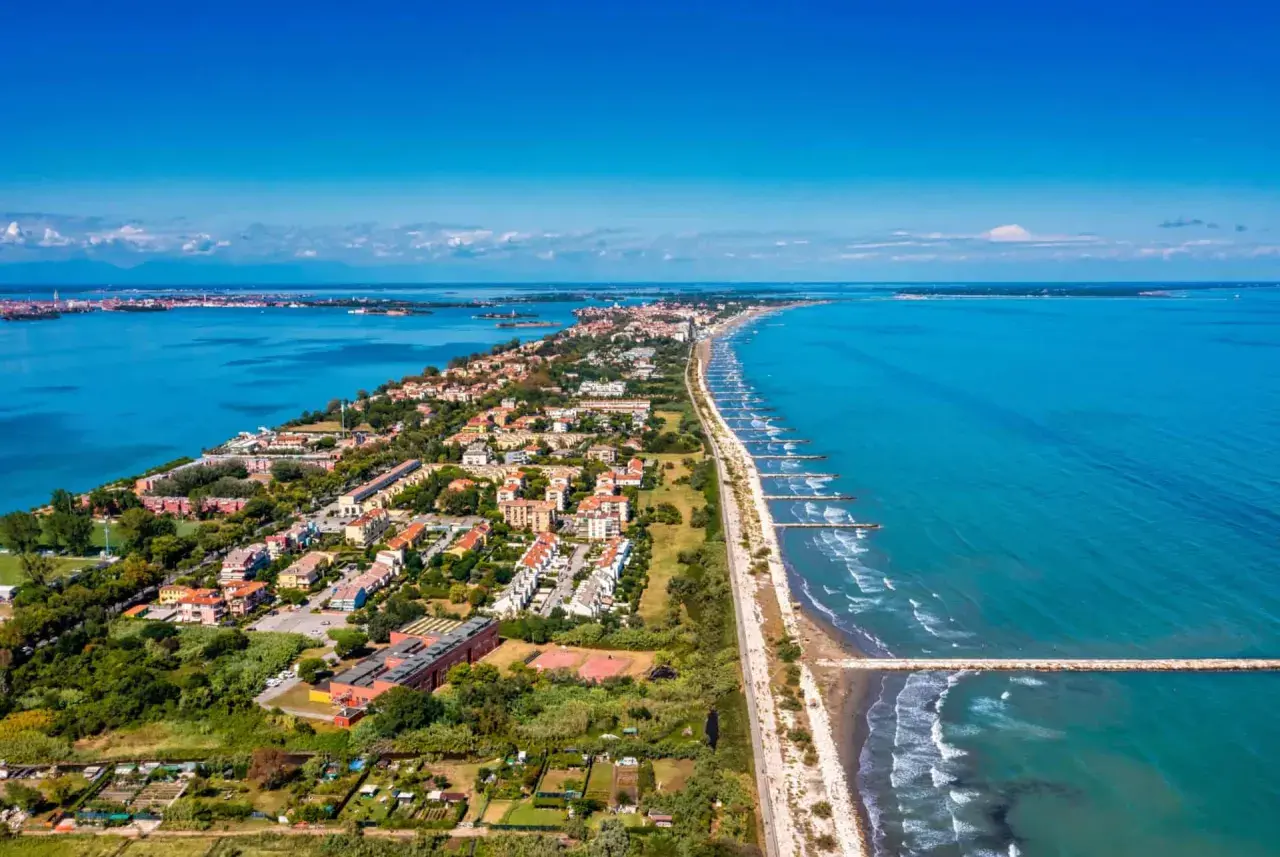 Vue aérienne du Lido di Venezia, une longue bande de terre bordée par la mer Adriatique et la lagune, avec des maisons, des arbres et des digues.