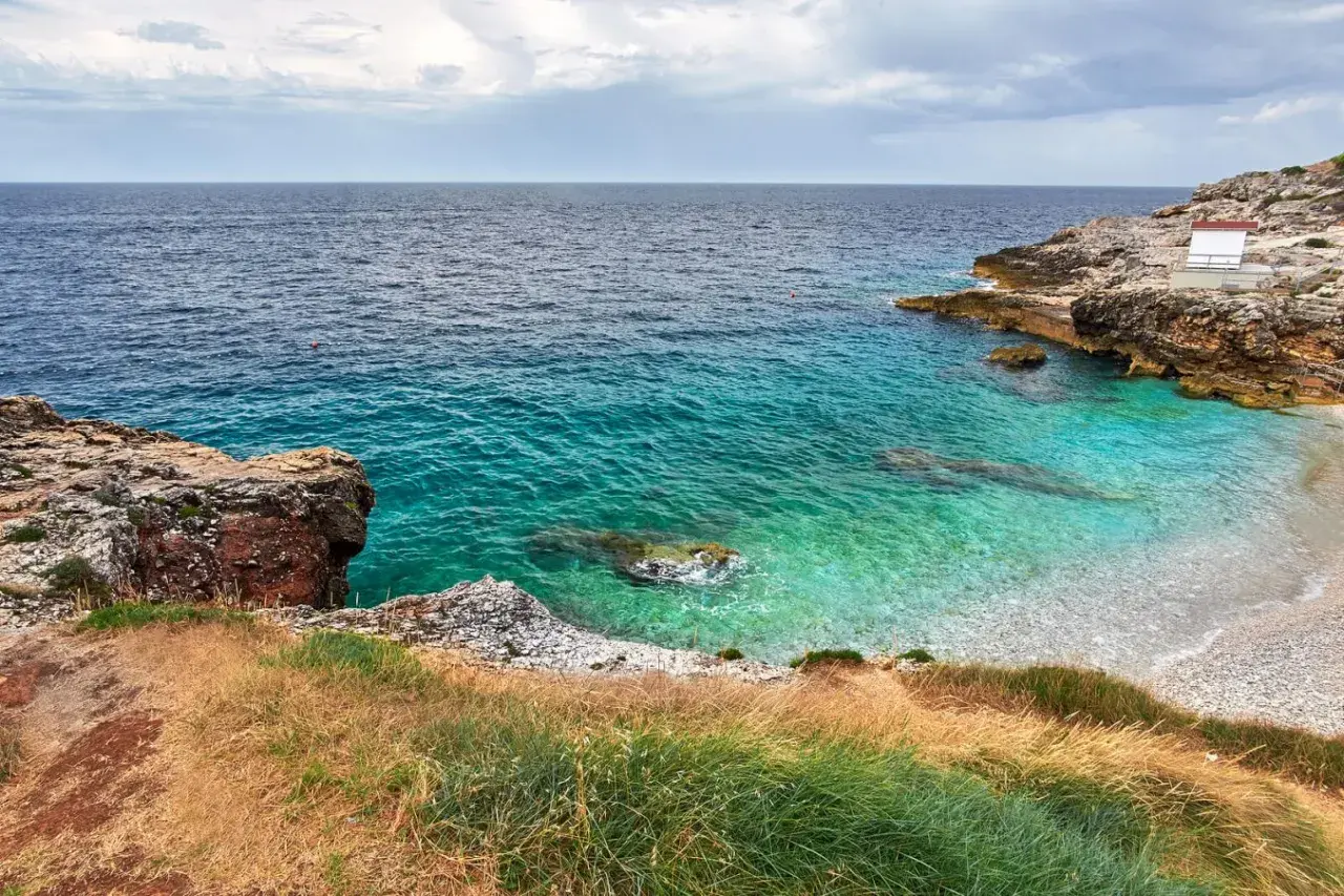 Vue d'une crique aux eaux turquoise cristallines, typique d'une plage de Pula en Sardaigne. Rochers et végétation méditerranéenne bordent la petite plage de galets.