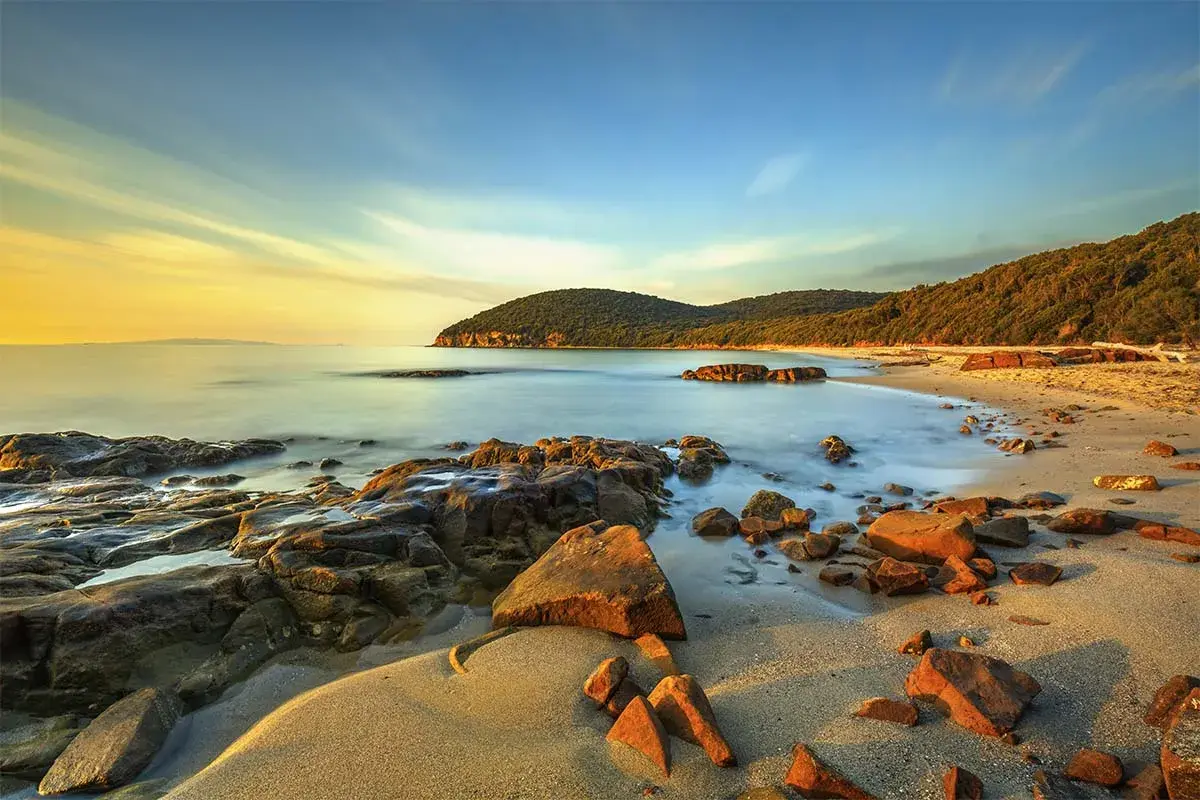 Coucher de soleil sur une plage rocheuse en Maremma, Italie. L'eau calme caresse le sable doré et les rochers ocre sous un ciel aux teintes chaudes.