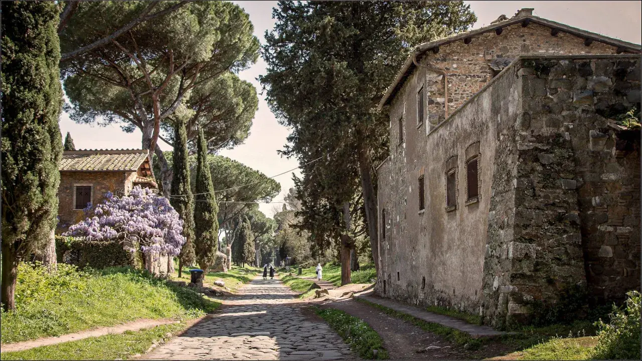 Chemin pavé bordé de cyprès et de pins, avec des maisons anciennes et des glycines en fleurs. Un air de via antica Roma.