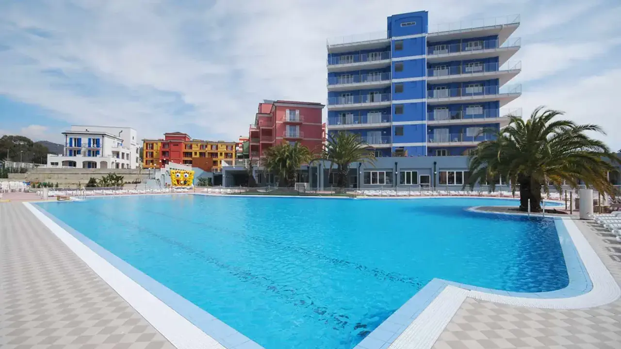 Vaste piscine bleue devant des bâtiments colorés et un grand gonflable de Bob l'éponge, au village ai pozzi.