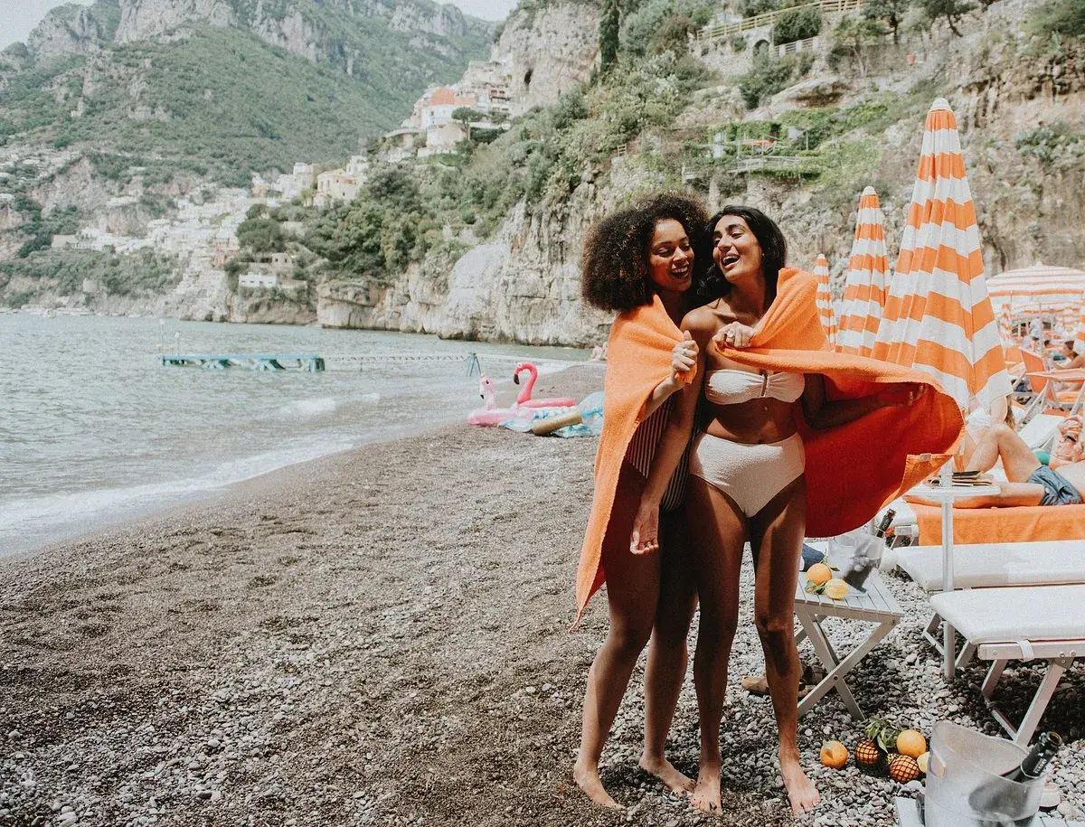 Deux amies rient sur une plage italienne, partageant une serviette orange. Leurs vacances en Italie en famille au bord de la mer sont idylliques.
