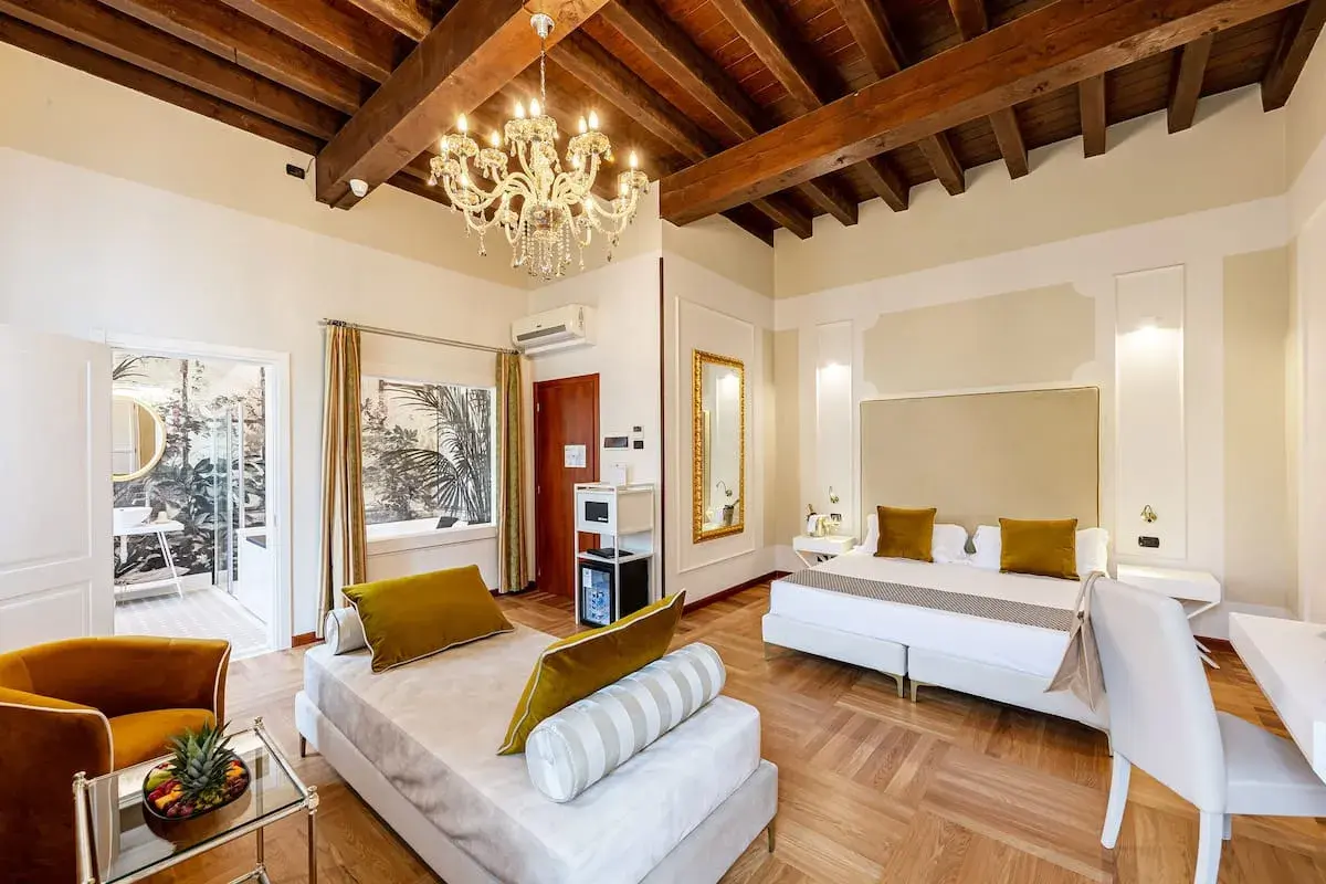 Suite luxueuse au **hotel Cariola Caprino Veronese** avec lit king-size, canapé, lustre et vue sur jardin tropical.