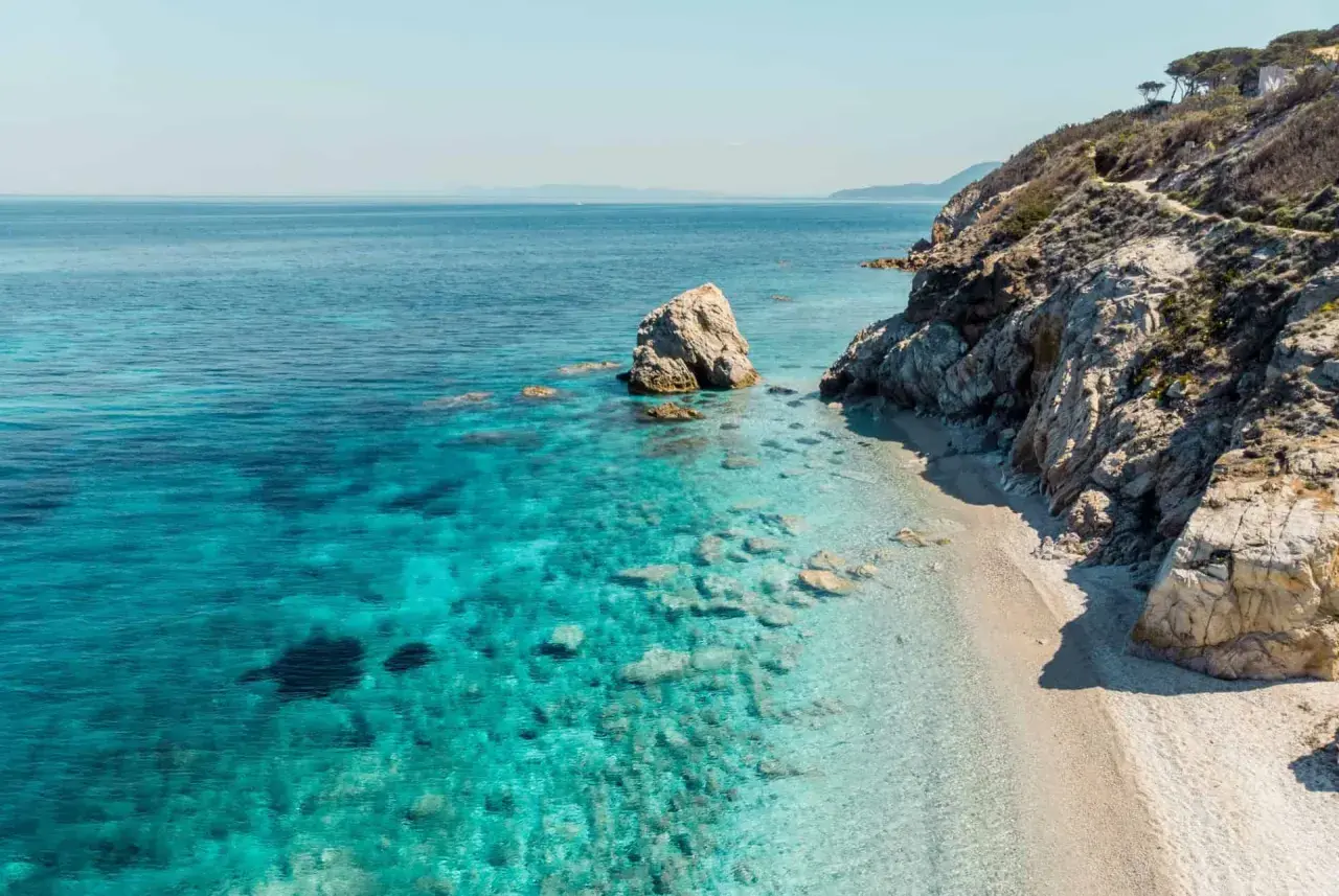 Plage de sable fin bordée par des eaux turquoise cristallines et des falaises rocheuses, typique des iles toscanes. Un rocher solitaire émerge de la mer.