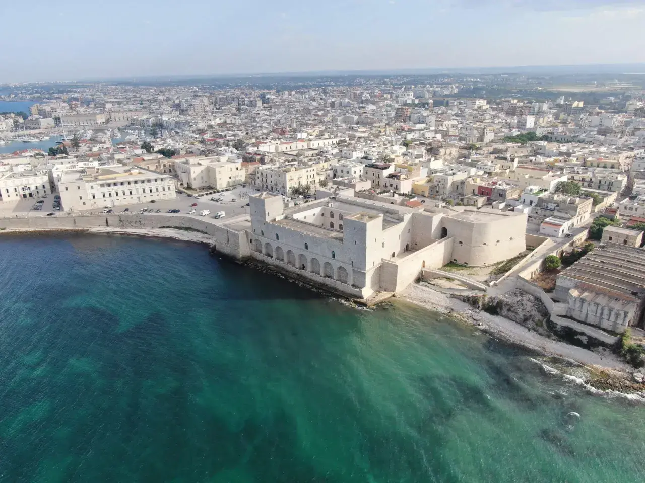 Vue aérienne du château de Trani, une forteresse médiévale sur la côte, avec la ville en arrière-plan et la mer d'un bleu-vert éclatant.