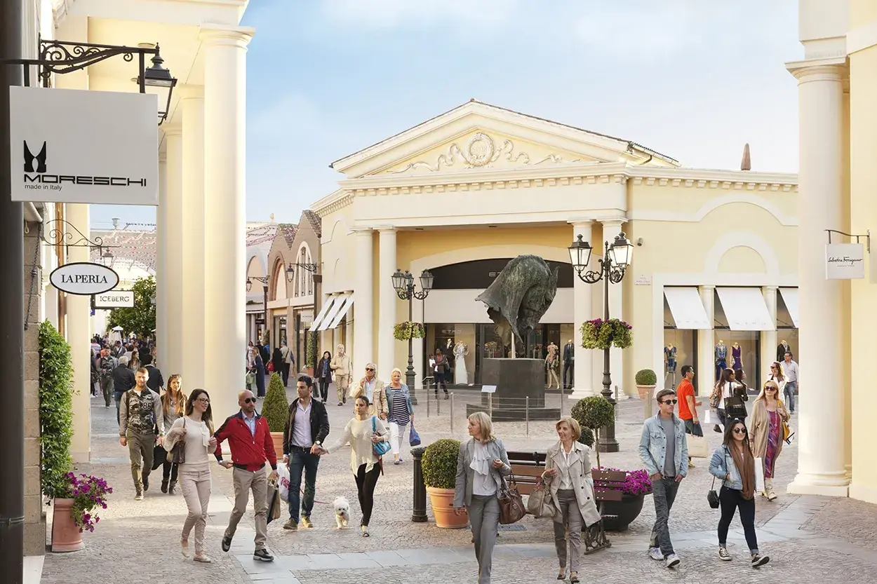 Des promeneurs profitent d'une journée ensoleillée au Castel Romano Outlet, admirant les vitrines et l'art.