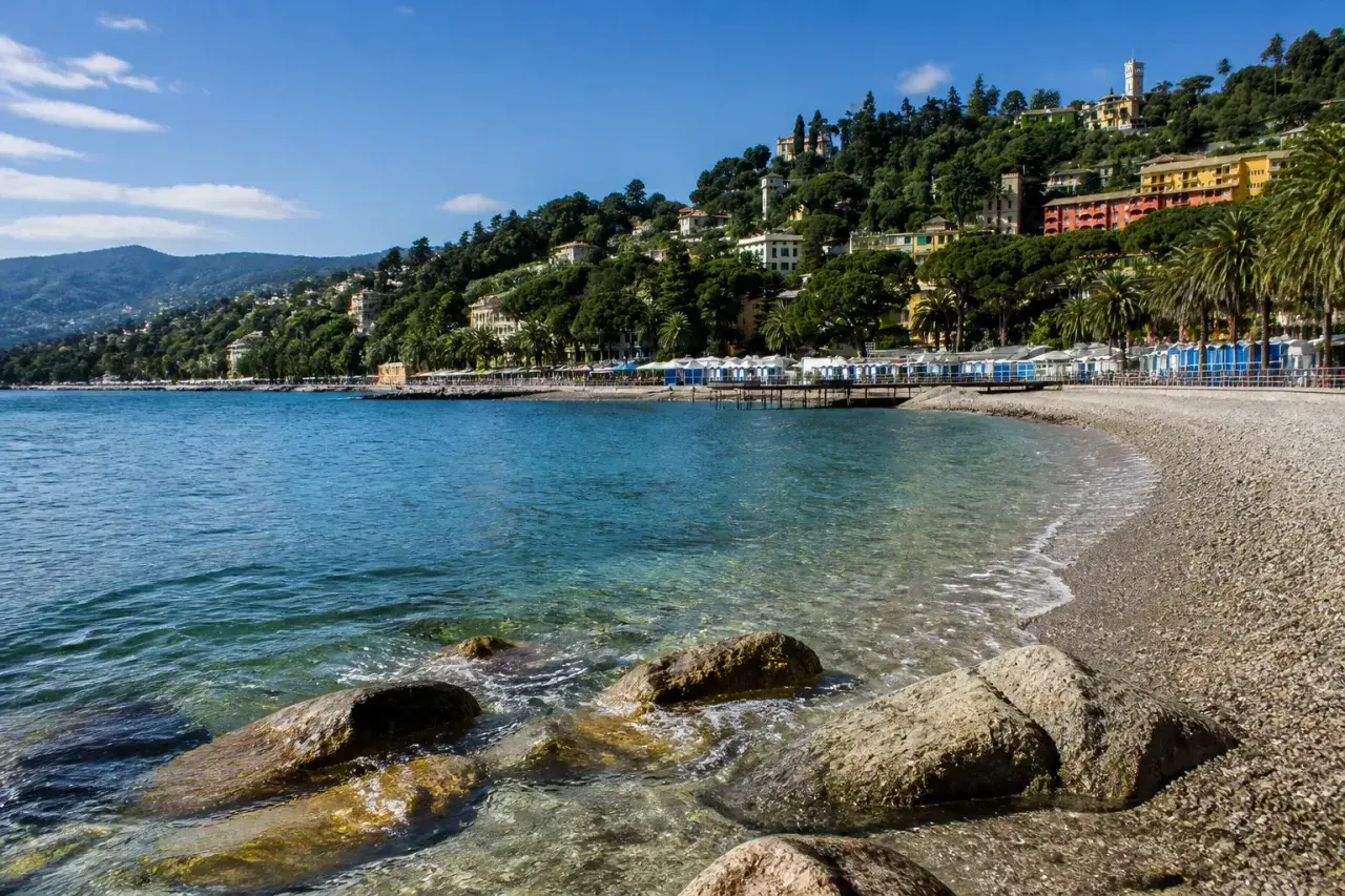 Plage de galets à Rapallo, avec des cabines bleues et blanches alignées, des maisons colorées sur la colline verdoyante et une mer bleue étincelante.