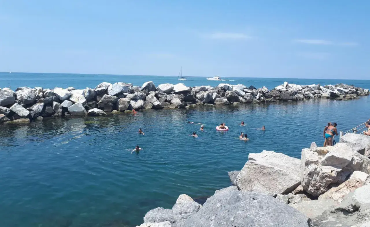 Baignade estivale dans la marina de Pise, avec des rochers formant une digue et des baigneurs profitant du soleil.