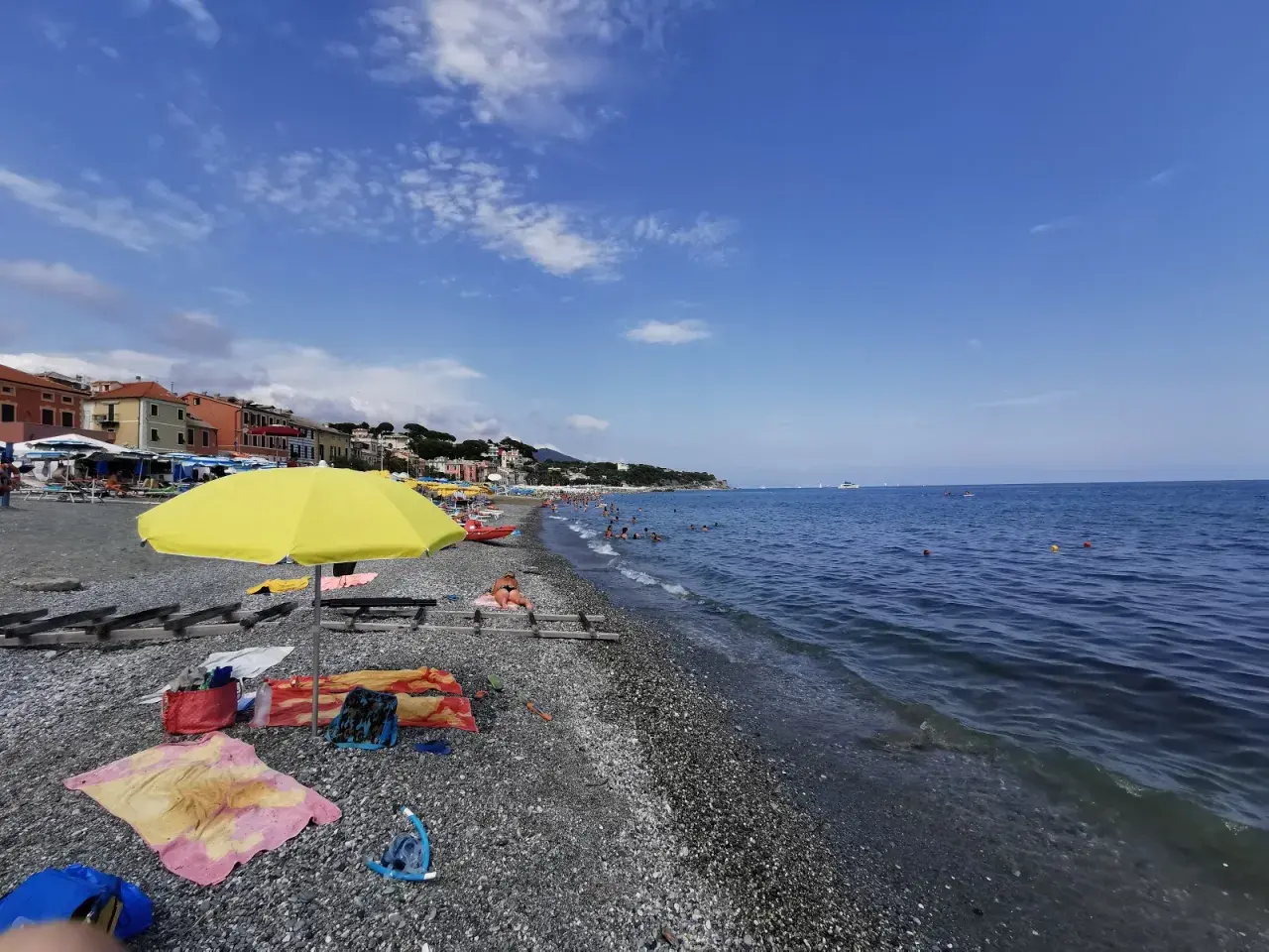 Plage de galets animée, avec des parasols, des serviettes et des baigneurs profitant du soleil. La mer bleue s'étend à l'horizon, avec des bateaux au loin. Un aperçu de la vie sur la côte ligure.