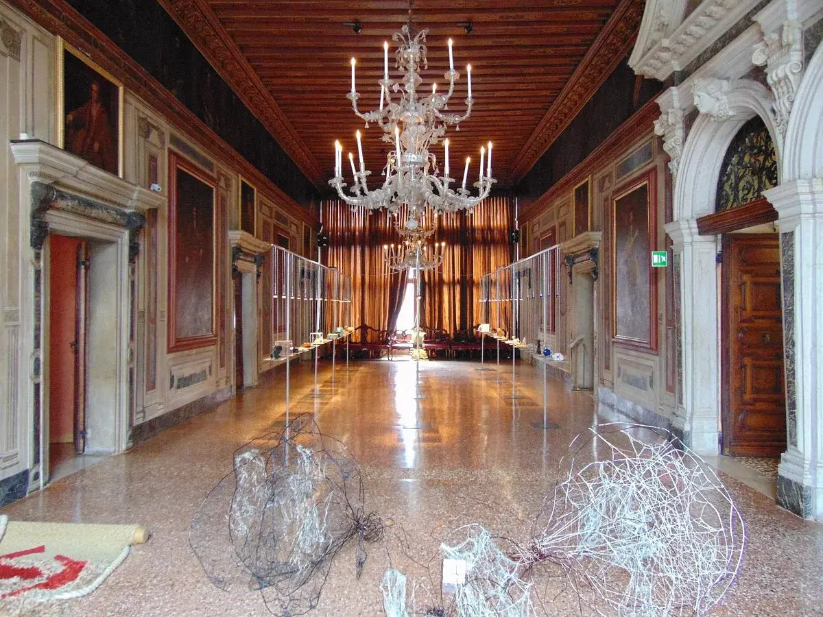 Intérieur du Palazzo Mocenigo, un long couloir orné de tableaux, avec un lustre imposant et des sculptures modernes au sol.