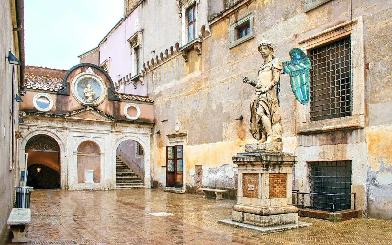 Intérieur du **château Saint-Ange** : une cour pavée et humide, une statue d'ange imposante, une arche menant à un escalier et des fenêtres grillagées.